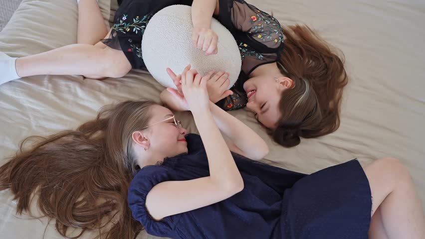 Two friends smiling together on beige bed in sunlit bedroom, holding hands and talking. Smiling, bed, friendship, sunlit, talking, hands.