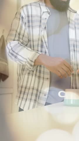 Vertical video: Bearded man at counter, right hand reaching mug, stirring, bokeh showing drink prep. Kitchen, domestic, cozy, plaid, cup, towel, vertical video
