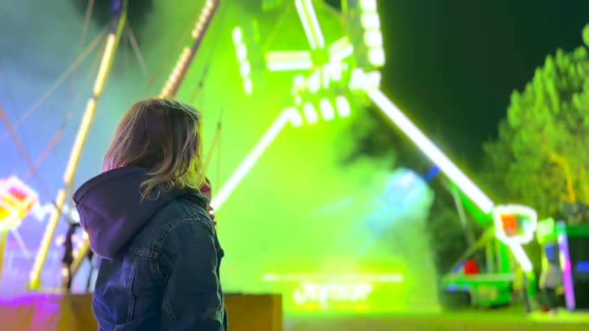 Young child with long hair watching a large amusement park attraction at night, captivated by the bright, changing neon lights and the thrilling atmosphere of the fairground during a festival