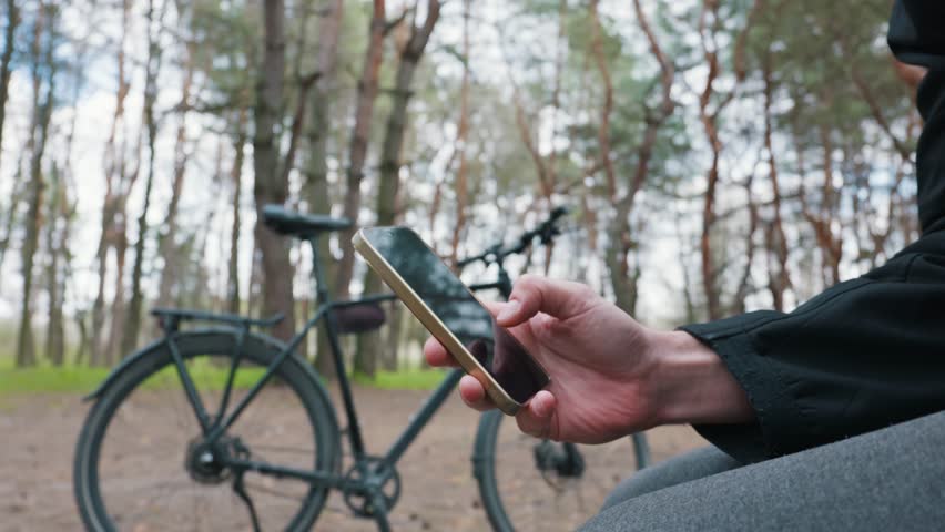 Man scrolls on a smartphone screen while taking a break during a bike ride in a pine forest. Represents connectivity and navigation during outdoor recreation.