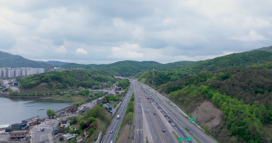 A cinematic aerial view of Baegun Lake in Uiwang, Gyeonggido, South Korea. Calm water is surrounded by lush green mountains and nearby residential areas, with a highway in the foreground showing light traffic. Ideal for themes of nature, urban harmony, travel, environment, and peaceful landscapes near metropolitan areas.