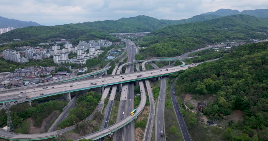 A cinematic aerial view of Baegun Lake in Uiwang, Gyeonggido, South Korea. Calm water is surrounded by lush green mountains and nearby residential areas, with a highway in the foreground showing light traffic. Ideal for themes of nature, urban harmony, travel, environment, and peaceful landscapes near metropolitan areas.