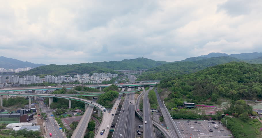 A cinematic aerial view of Baegun Lake in Uiwang, Gyeonggido, South Korea. Calm water is surrounded by lush green mountains and nearby residential areas, with a highway in the foreground showing light traffic. Ideal for themes of nature, urban harmony, travel, environment, and peaceful landscapes near metropolitan areas.