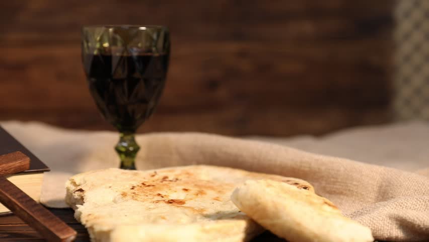 Sacrament of Communion. Glass of red wine, flatbread, cross, rosary and Bible on wooden table, closeup. Camera moving left