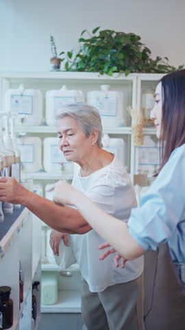 Senior Asian woman smells a wooden tester stick to check the scent of a bulk liquid product while shopping at a local eco-friendly zero waste refill store with a clerk.