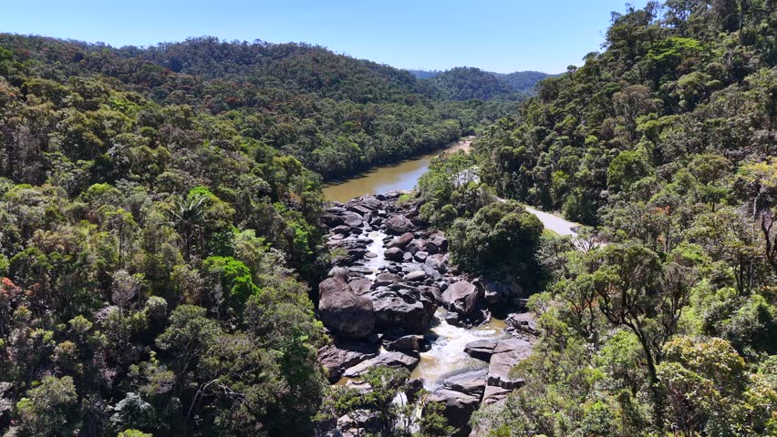 Aerial view shows winding river through lush green forest. River flows over rocks, creating small waterfalls. Road curves beside the river, following valley terrain