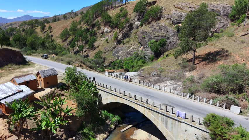 Scenic road winds through mountainous terrain under bright blue skies. Concrete bridge spans a rocky gorge with people walking across. Small buildings and greenery line the road