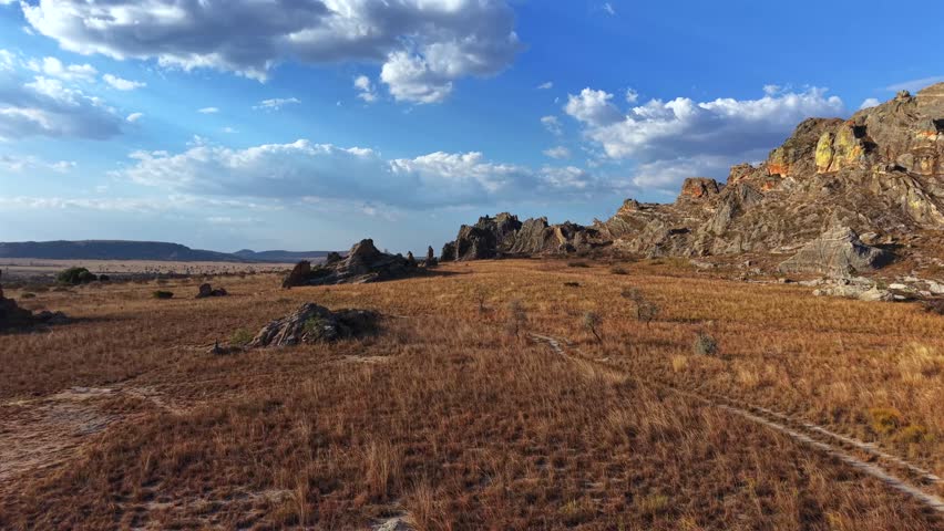 Aerial view of rocky terrain under bright blue sky. Dry grass covers the ground in golden hues. Clouds drift slowly across the horizon. Rocky outcrops rise sharply from the earth