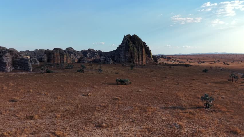 Desert landscape with rocky outcrop in dry, flat terrain. Sky clear with few clouds, sunlight bright. Sparse, low vegetation covers ground. No people or animals visible