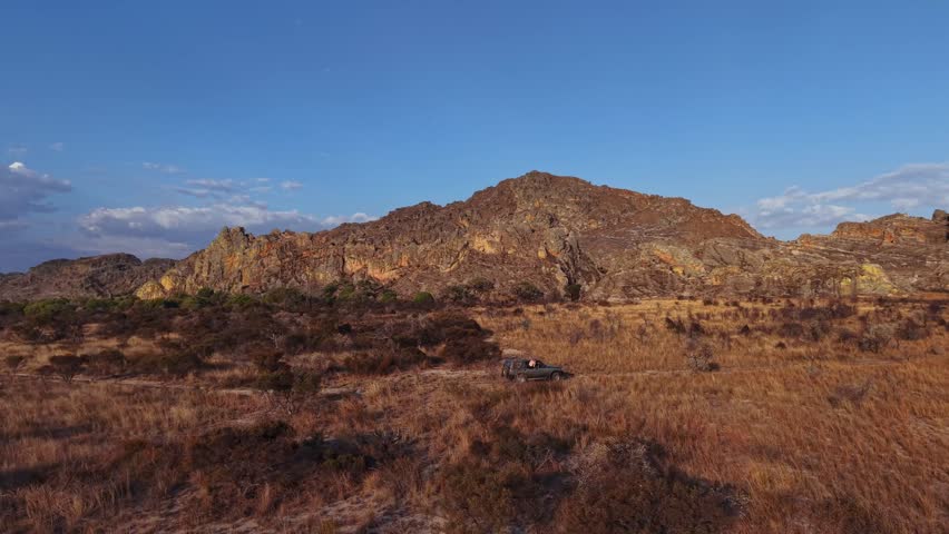 Desert landscape with rugged hills and dry grass under clear blue sky. Sunlight casts warm glow on rocky terrain. Dirt trail winds through barren land. Scene evokes solitude, adventure