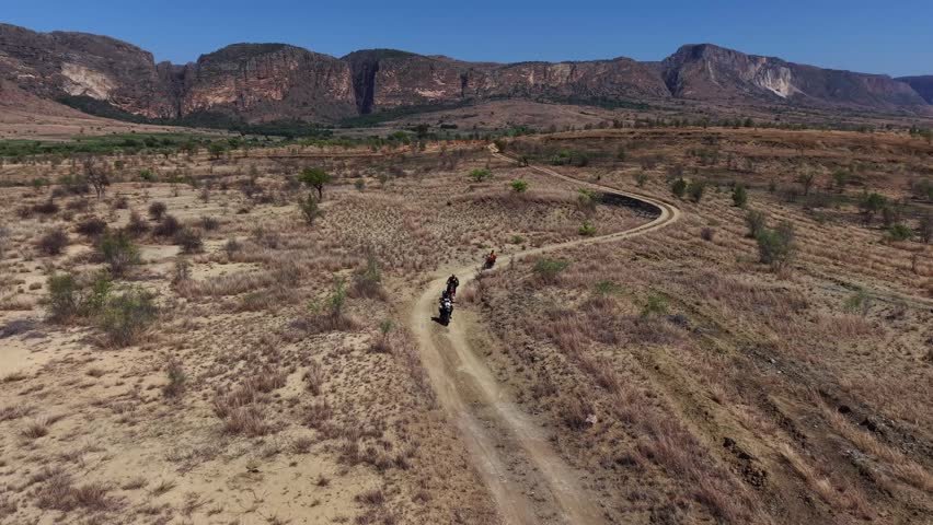 Aerial view of a dry, arid landscape with winding dirt roads. People ride horses along the trail, moving calmly through sparse vegetation. The distant mountains rise sharply under a clear blue sky