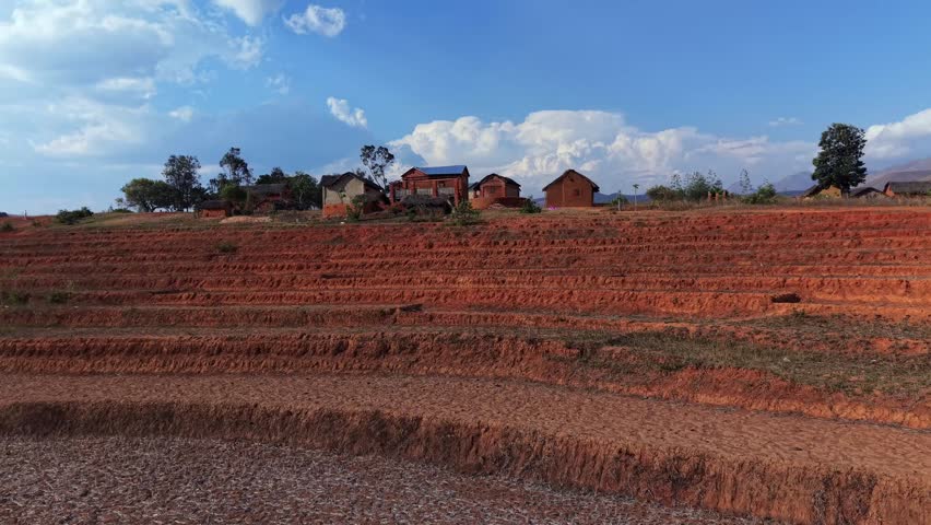 Red clay terraces stretch across a rural landscape. Small houses dot the hillsides beneath a bright blue sky. Mountains frame the distant horizon. No people visible