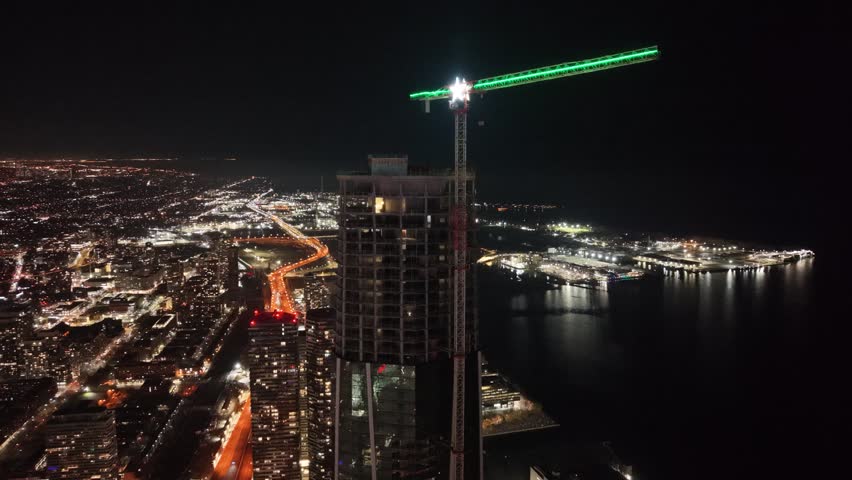 A cinematic clockwise aerial drone orbit around a skyscraper under construction on Toronto’s waterfront, revealing the Gardiner Expressway, illuminated crane star, and Lake Ontario at night