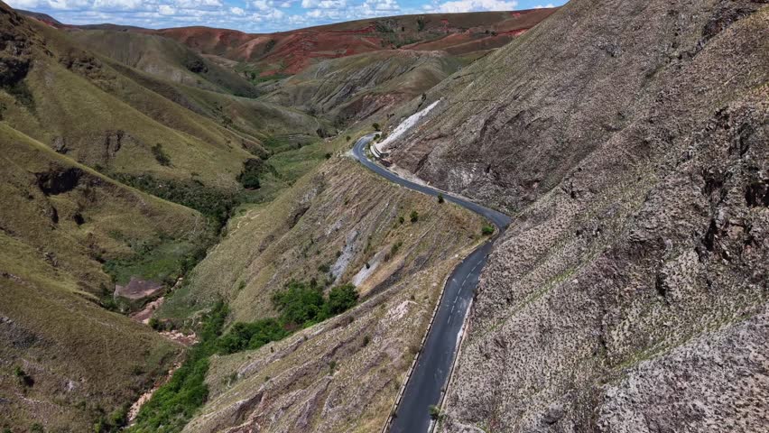 Winding mountain road curves through rocky valleys under clear blue skies. Vehicle travels the narrow path, navigating steep slopes. The terrain features rugged cliffs and sparse greenery