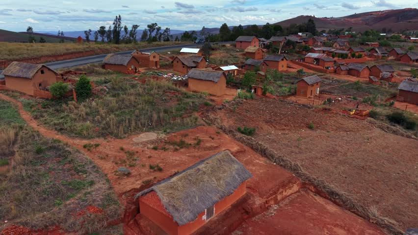 Aerial view of rural village nestled among red hills. Houses built from local clay and wood line winding roads. Scattered trees dot dry fields under soft sky. No people visible