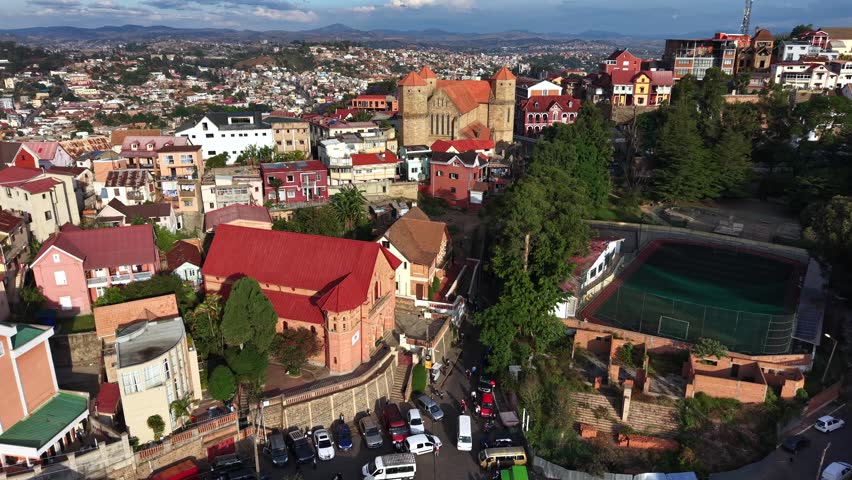 Aerial view of colorful hillside town with historic church and winding streets. Red roofs contrast with green trees and distant mountains. Quiet atmosphere, no visible people