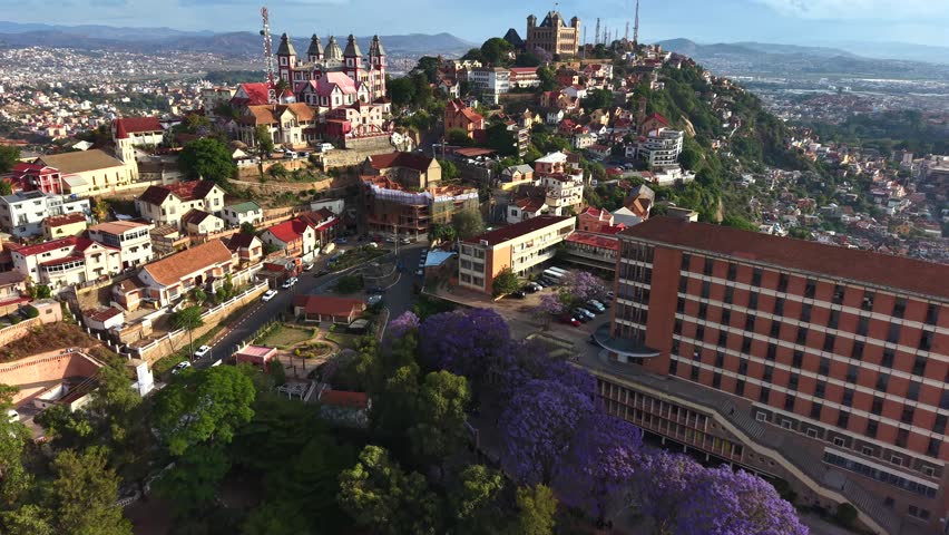 Aerial views capture vibrant hillside town with historic church and colorful homes. Purple flowering trees add vivid contrast to red rooftops. City sprawls below under cloudy skies