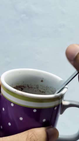 A hand holds a spoon with used coffee grounds over a purple polka-dotted mug 