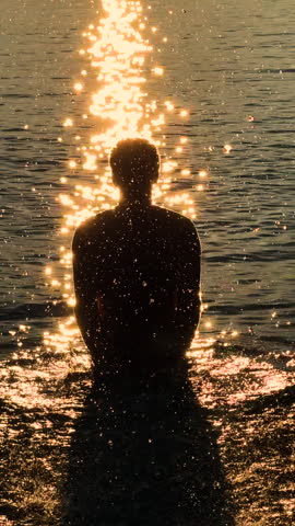VERTICAL, LENS FLARE, SILHOUETTE, SLOW MOTION: Backlit man stands in shallow sea water with raised arms, sending sparkling droplets into the air. Playful guy splashing sea in golden sunset light.