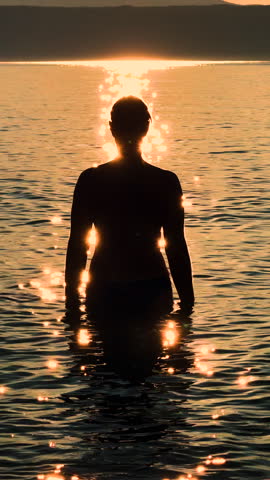 VERTICAL, LENS FLARE, SILHOUETTE, SLOW MOTION: Backlit young woman stands in shallow sea with raised arms, tossing sparkling droplets into the air. Playful lady splashing water in golden sunset light.