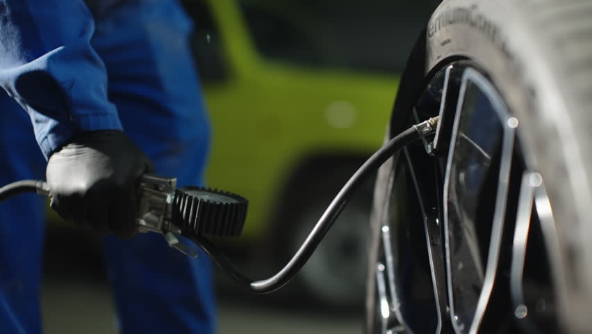 Close-up of a mechanic inflating a car tire with a pressure gauge and air hose, checking wheel pressure during automotive maintenance in a garage, wearing gloves and coveralls