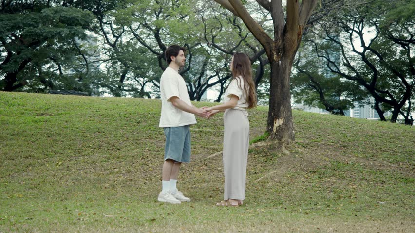 Medium shot portrays affectionate duo locked in loving hug beside rough textured tree base surrounded by vibrant grass patches winding park trails tall surrounding trees and hazy horizon