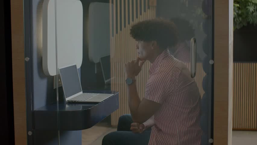 Adult male sitting in pod, viewing laptop for tech while blue waveform sweeping glass driving focus. Privacy, booth, coworking, concentration, monitor, computer, minimal
