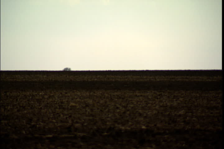 Long, slow pullback and pan over flat horizon from empty field to dirt road in Russell, Kansas.