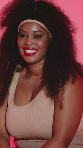 Vertical video: Smiling woman camera starting, leaning, easing back in pink set beige tank headband. Studio, backdrop, portrait, jewelry, lipstick, earrings, vertical video