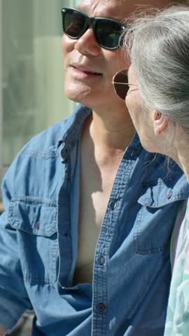 Vertical video: Opening mouth aged man dark shades blue shirt sharing by glass door woman listening. Senior, couple, sunglasses, window, patio, outdoor, vertical video