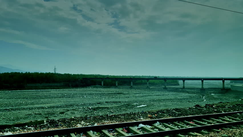 A moving train view in India showing railway tracks in foreground with a wide dry riverbed and a long bridge stretching across under an overcast sky.