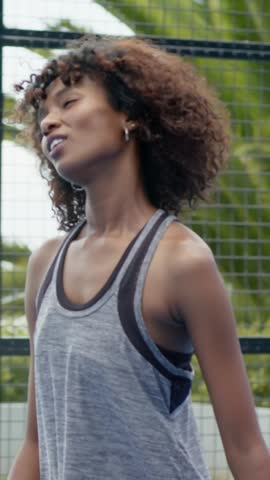 Vertical video: Woman grasping hand coming left, greeting by mesh fence, gray tank, black sportsbra. Female, athletic, outdoors, enclosure, greenery, gesture, vertical video