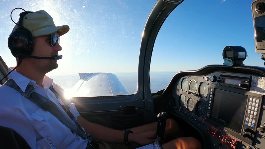 Pilot Flying Small Aircraft Cockpit with Avionics Dashboard and Clear Blue Sky Horizon. Young Male Pilot in Aviation Headset Controls Light Plane Cockpit During Daytime Flight