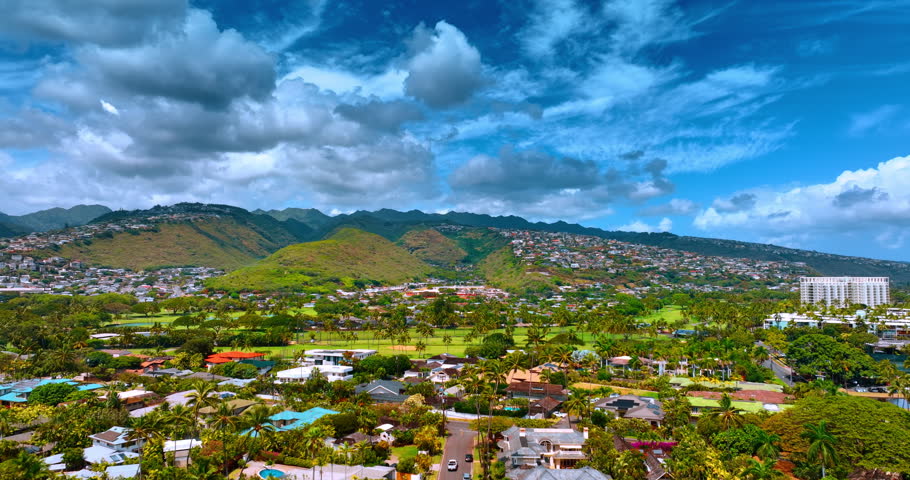 Villas and palm trees in Honolulu, Hawaii. Drone footage over the scenery approaching golf course. Cloudy sky above the mountains. Aerial view.