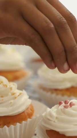 Vertical video: Hovering kid hand decorating cupcakes, pink pearls, gold dragee on rack, copy space. Child, frosting, sprinkles, commercial, closeup, bakery, vertical video