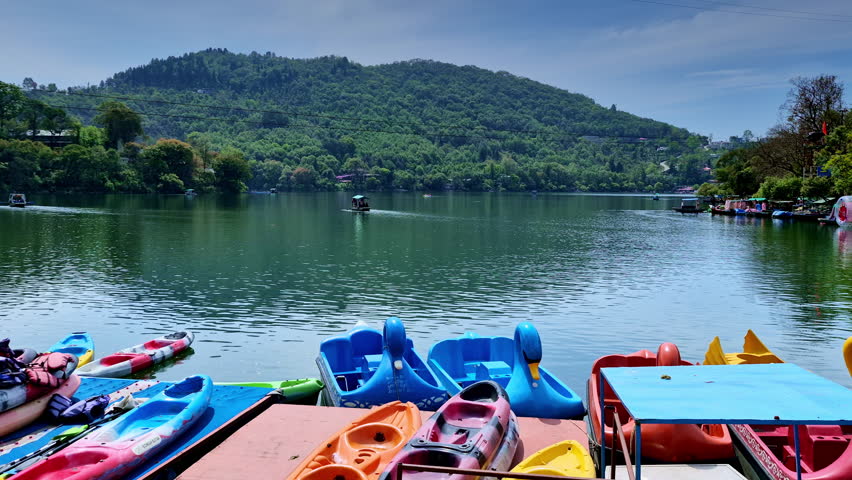 A serene lakeside view with colorful kayaks and swan-shaped paddle boats resting at dock, overlooking a wide calm lake with a lone boat drifting toward a lush green hillside under a clear sky.
