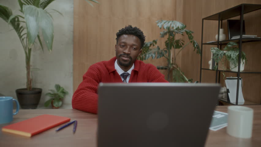 Young Black businessman sitting at desk in cozy office, smiling and talking to laptop webcam during video call or online meeting
