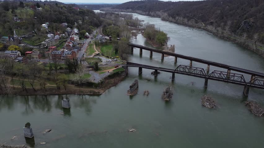 Lower Town Harpers Ferry sits between Potomac Shenandoah Rivers beneath Maryland Heights rail crossing. Brick blocks, metal roofs, soft spring canopies, river sheen define textures in WV. USA