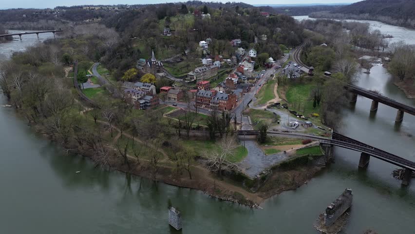Harpers Ferry riverfront curves along Potomac beside rail bridge narrow historic streets below wooded ridge. Mixed roofing, early leaf-out, warm hillside tones shape textures in WV. USA