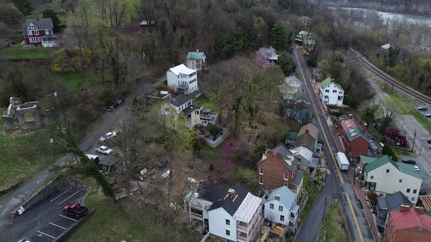 Harpers Ferry hillside homes line narrow streets descending toward river corridor rail alignment. Varied roof materials, muted spring canopies, soft valley haze define textures in WV. USA