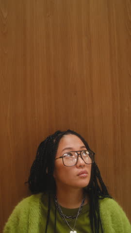 Portrait of young Asian woman with long braids and trendy oversized glasses, wearing bright lime green cardigan, looking at camera against minimalist wood wall. Vertical clip