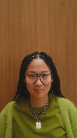 Stylish young Asian woman with braided hair and glasses, wearing casual green outfit, looking at camera and smiling against wooden wall background. Vertical shot, video portrait