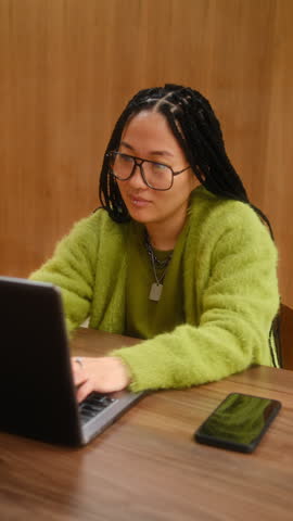Focused young Asian woman with braids and glasses, wearing lime green cardigan, working on laptop at wooden desk in modern minimalist office. Vertical shot