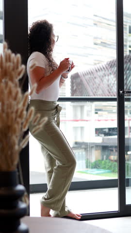 Pensive barefoot woman with curly hair standing by a large glass window, looking at the city view while drinking a warm beverage from a transparent mug on a peaceful morning at home