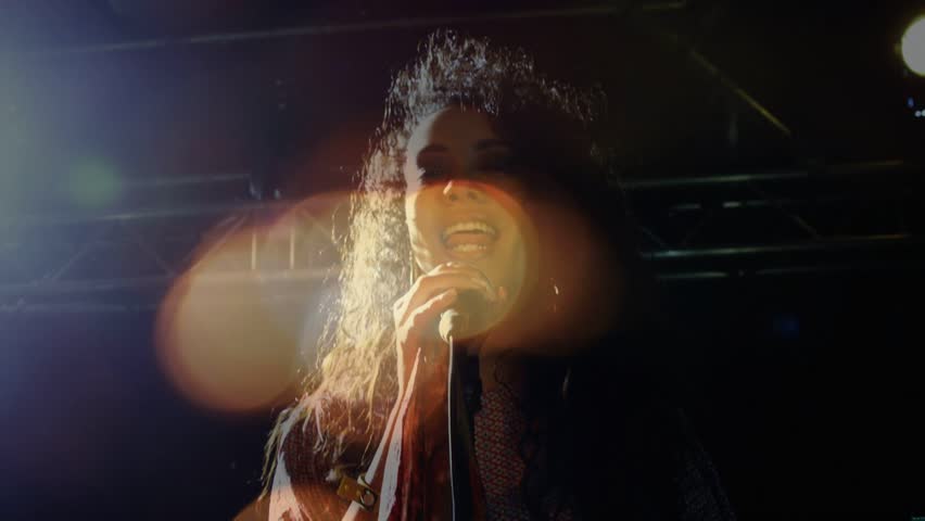 Female singer opening note into mic, causing concert lights, lens flares fanning hair to wow crowd. Performer, microphone, backlit, stage, editorial, motion, energy