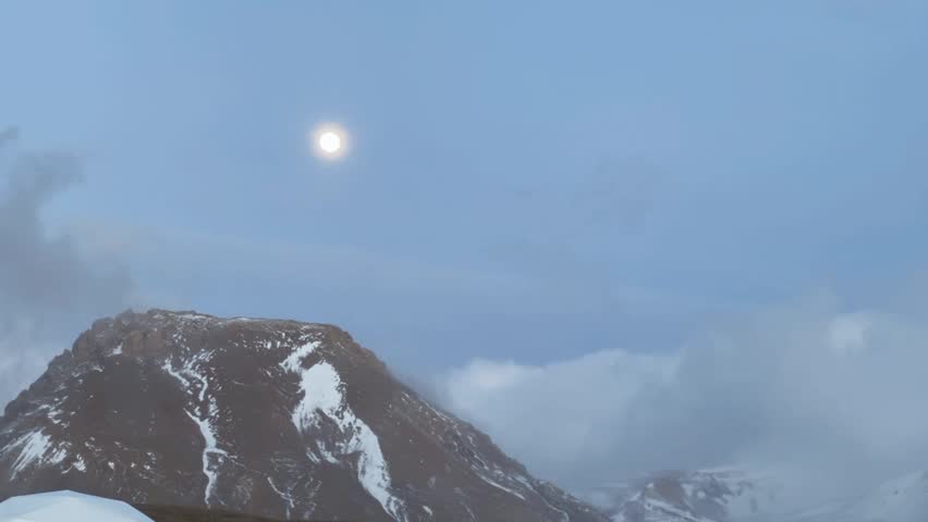 A cinematic shot of a bright moon in the twilight sky over a rugged snow-dusted mountain peak in Tibet China.