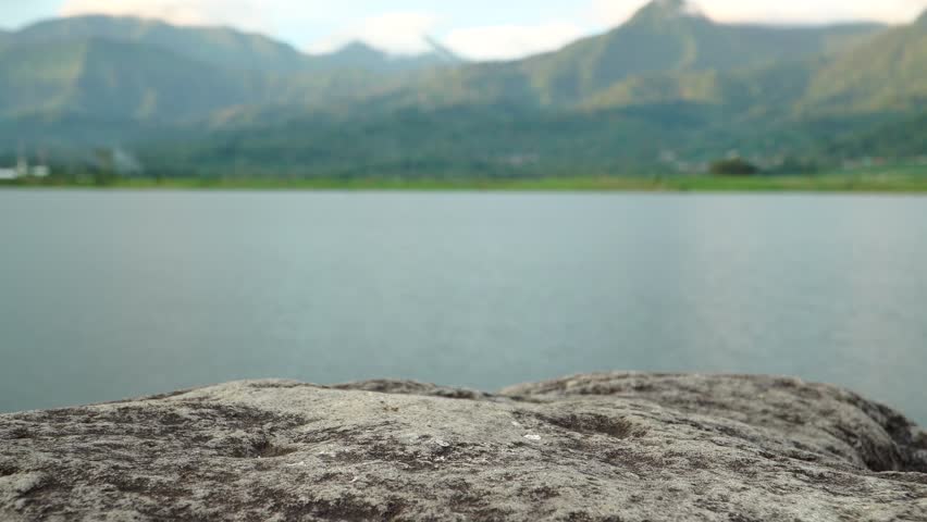 Large textured rock surface in the foreground acting as an empty display stage, set against a blurred background of a wide lake and distant green mountains in the afternoon light.