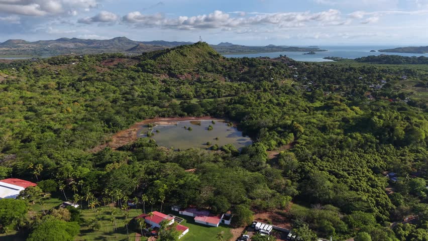 Aerial view shows lush green forests surrounding a calm pond. Mountains stretch into distance under bright blue sky. Small structures with red roofs dot the landscape