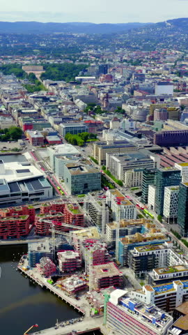 Vertical video. Oslo, Norway. The Oslo Opera House dominates the waterfront with its angular white Carrara marble and granite surfaces that appear to emerge directly from the Oslo Fjord. Designed by Snohetta and opened in 2008, the building features a publicly accessible sloping roof that creates a seamless transition from the city plaza to the water, embodying the Scandinavian principle of allemannsretten, the right to roam. The surrounding Bjorvika district showcases contemporary high-rise architecture including the distinctive Barcode Project towers and the nearby Munch Museum, set against the backdrop of the Oslo Fjord and distant forested hills. The scene is captured in bright daylight with clear skies, highlighting the contrast between the pristine white opera house, the deep blue water, and the varied modern structures of the redeveloped harbor area. The aerial perspective reveals the full integration of cultural architecture with urban waterfront renewal. Aerial View. Rich colors