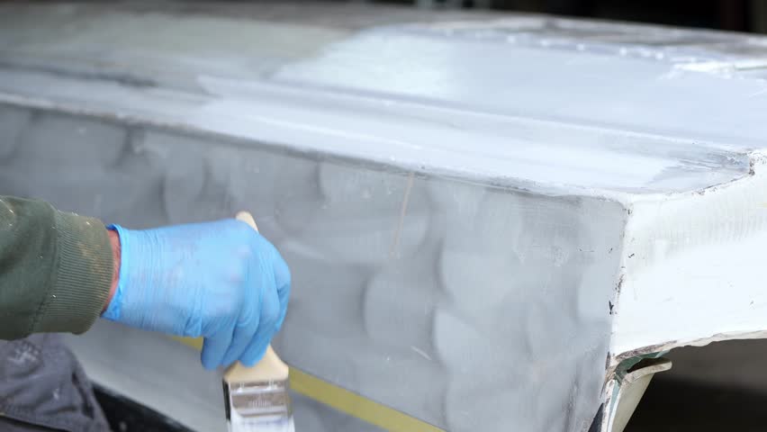 A person paints the underside of a small fiberglass boat with a brush, showing surface preparation and careful coating during a DIY marine restoration process.